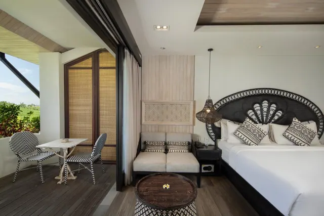A bedroom with a king bed under a woven black headboard, a cream armchair pair, and a balcony with striped chairs and a marble table overlooking green fields and trees.