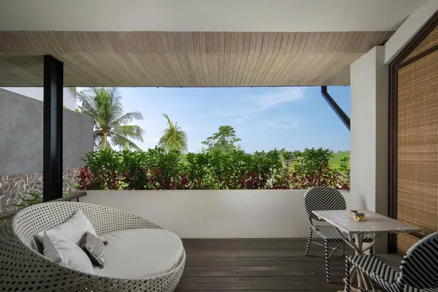 A covered balcony with a wicker daybed, cushions, two striped chairs and a wooden table, looking out over palm trees and green foliage.