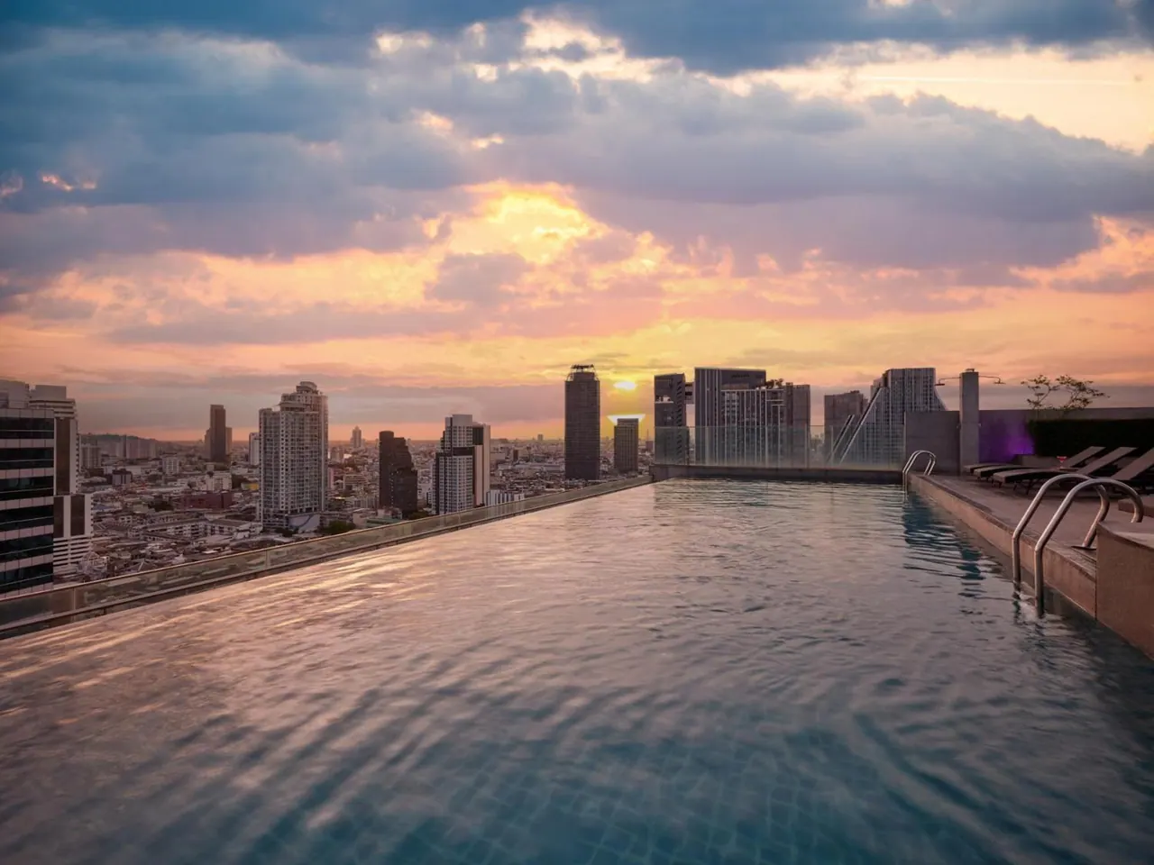 A rooftop infinity pool at sunset overlooks a city skyline, with sun loungers along the far edge.