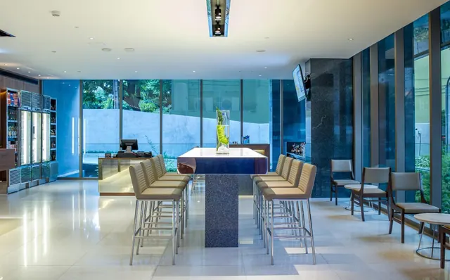 A lounge with a long high table, bar stools, armchairs, a reception counter and glass-door beverage fridges beside floor-to-ceiling windows that look out onto trees and a wall.