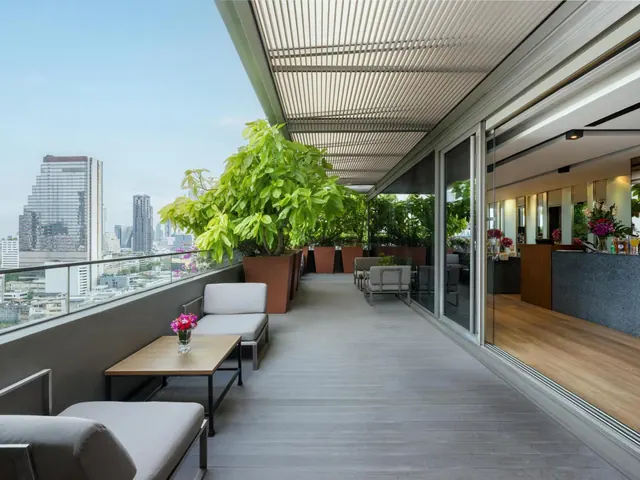 A covered terrace with lounge seating and potted trees overlooks a city skyline, with an indoor bar visible through sliding glass doors.