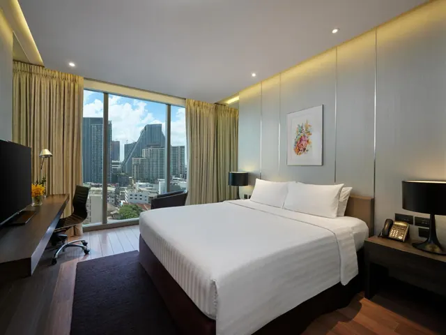 A bedroom with a king bed, a wooden work desk with a chair, a television, and floor-to-ceiling windows overlooking city high-rises.