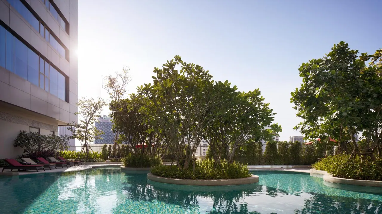 An outdoor pool with turquoise water surrounded by trees in circular planters, with pink sun loungers on the left beside a tall building.
