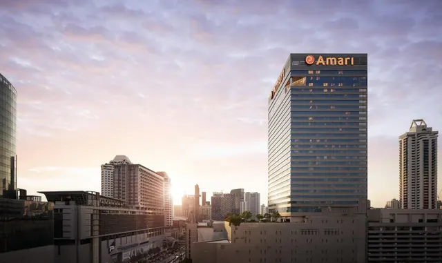 Exterior of a glass-fronted Amari hotel tower with its name on the roof, with other high-rise buildings and a sunset sky behind it.