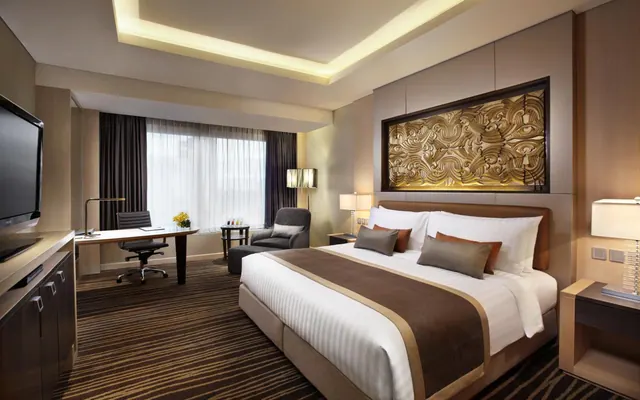 A king bed with white bedding and a carved gold wall panel sits beside a work desk, an armchair and a wall-mounted television in a hotel bedroom.