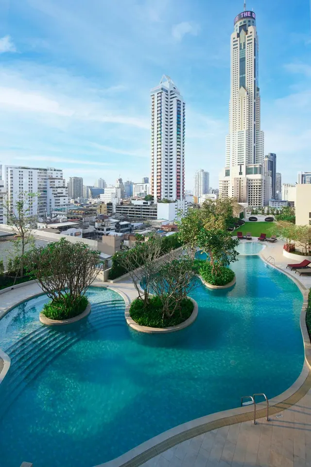 A large curved swimming pool with tree-planted islands, with high-rise towers and the Baiyoke Sky tower rising behind it under a blue sky.