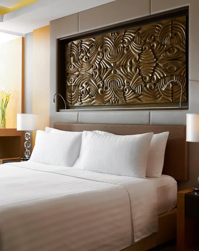 A made bed with white pillows and striped linen sits below a carved wooden wall panel, with a bedside lamp on each side.