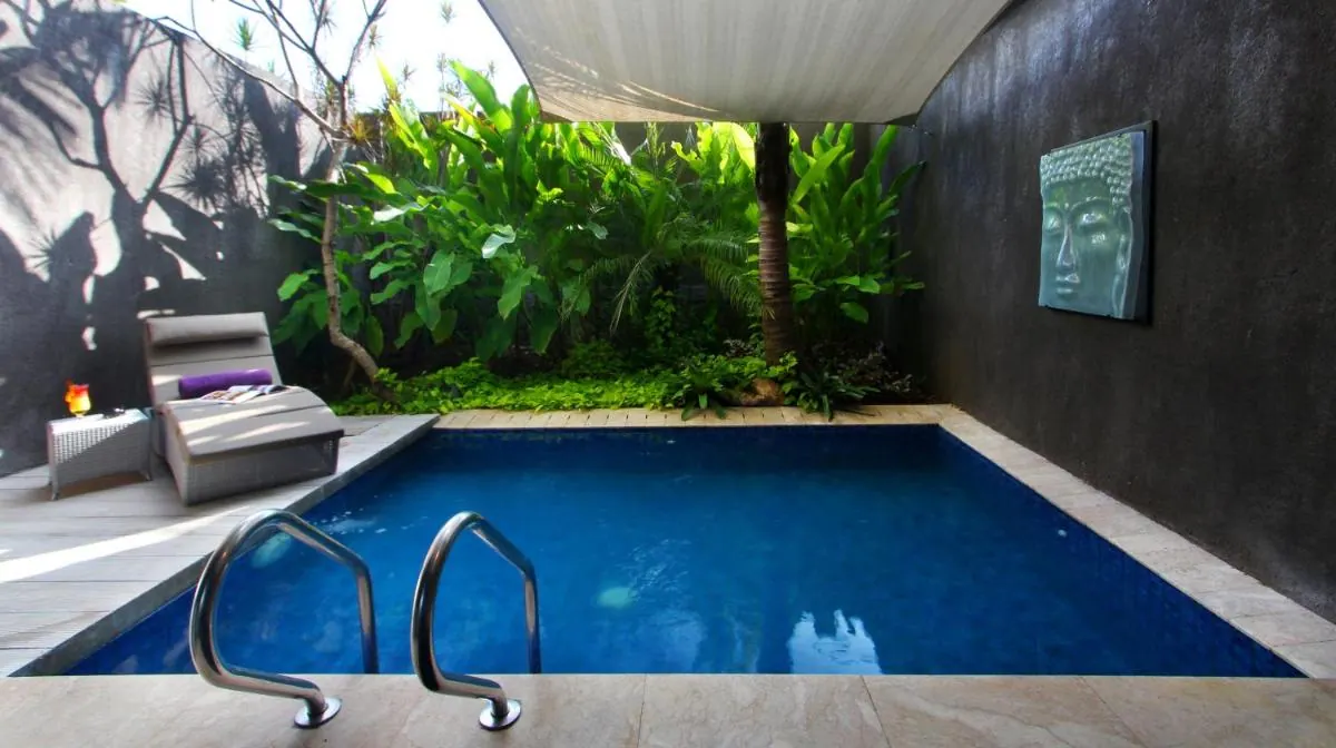 A small private plunge pool sits in an enclosed courtyard with a sun lounger, a steel pool ladder, a Buddha wall relief and tropical plants behind.