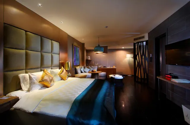 A bedroom with a large king bed under a tall padded headboard, a sofa and round table seating area, and a wall-mounted television.