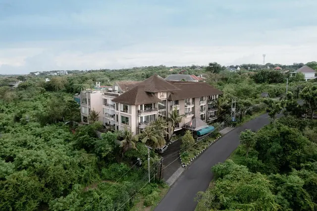 Aerial view of a multi-storey hotel building with brown pitched roofs and balconies, beside a paved road and surrounded by dense green trees and palms.