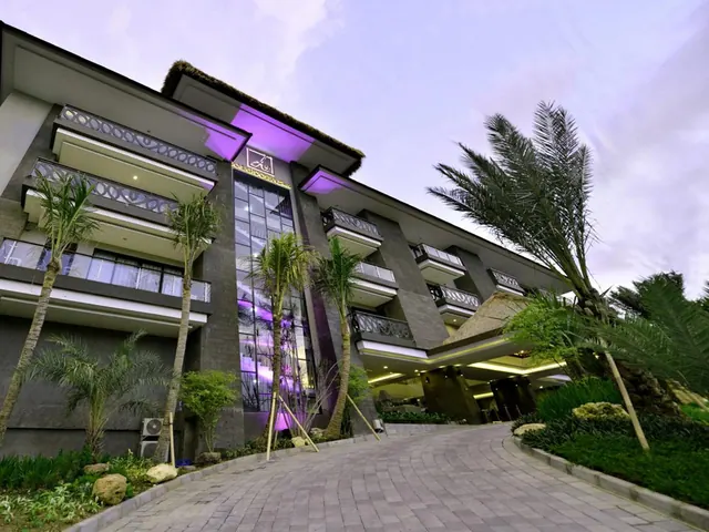 Low-angle dusk view of a multi-storey hotel building with purple uplighting, palm trees, and a paved driveway leading to the entrance.