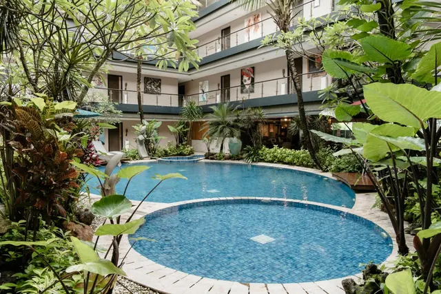 A courtyard swimming pool with a large oval basin and a small round basin is surrounded by tropical plants, with hotel balconies rising behind it.