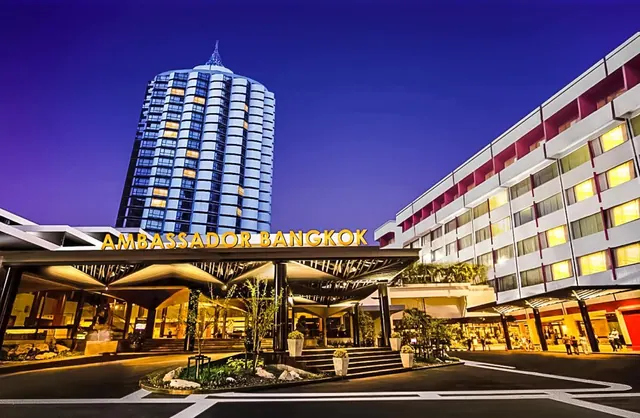 Twilight exterior of the Ambassador Bangkok hotel, with its illuminated name sign over the entrance canopy, a tall tower behind it and a wing extending to the right.