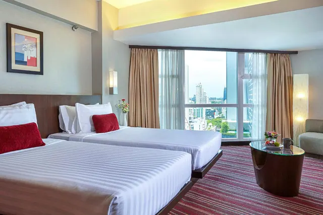 A bedroom with two beds dressed in white linen and red cushions, a seating area, and a large window overlooking city buildings.