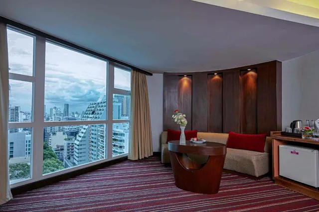 A living area with floor-to-ceiling windows looking over high-rise buildings, a curved sofa with red cushions, a round wooden table and a small fridge.