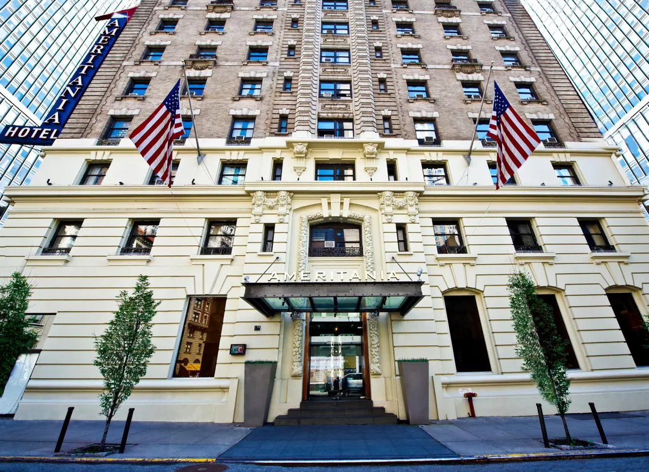 The front of the Ameritania hotel, with a cream facade, an entrance canopy, two American flags and a tall brick tower rising behind it.