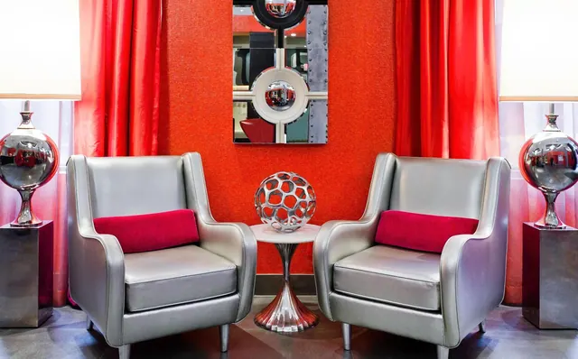Two silver armchairs with red cushions face each other beside a round side table with a metal sculpture, flanked by chrome lamps, with red curtains and a wall mirror behind.