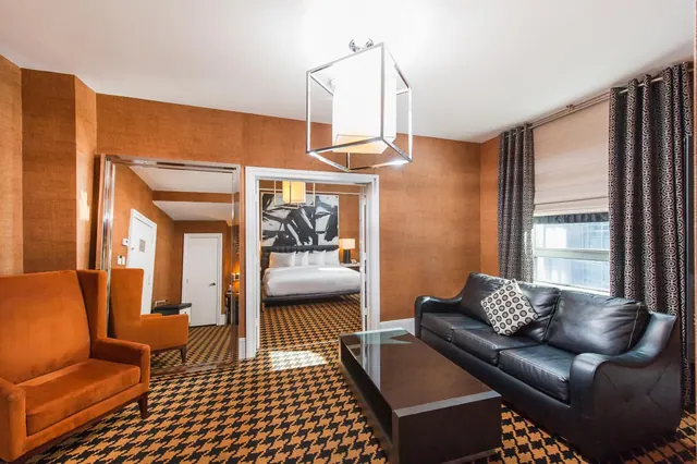 A living area with orange armchairs, a black leather sofa and a dark coffee table, with a bed visible through the open doorway.