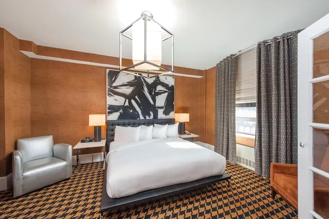 A bedroom with a king bed on a platform base, a black and white abstract painting above the headboard, two armchairs, and houndstooth carpet.