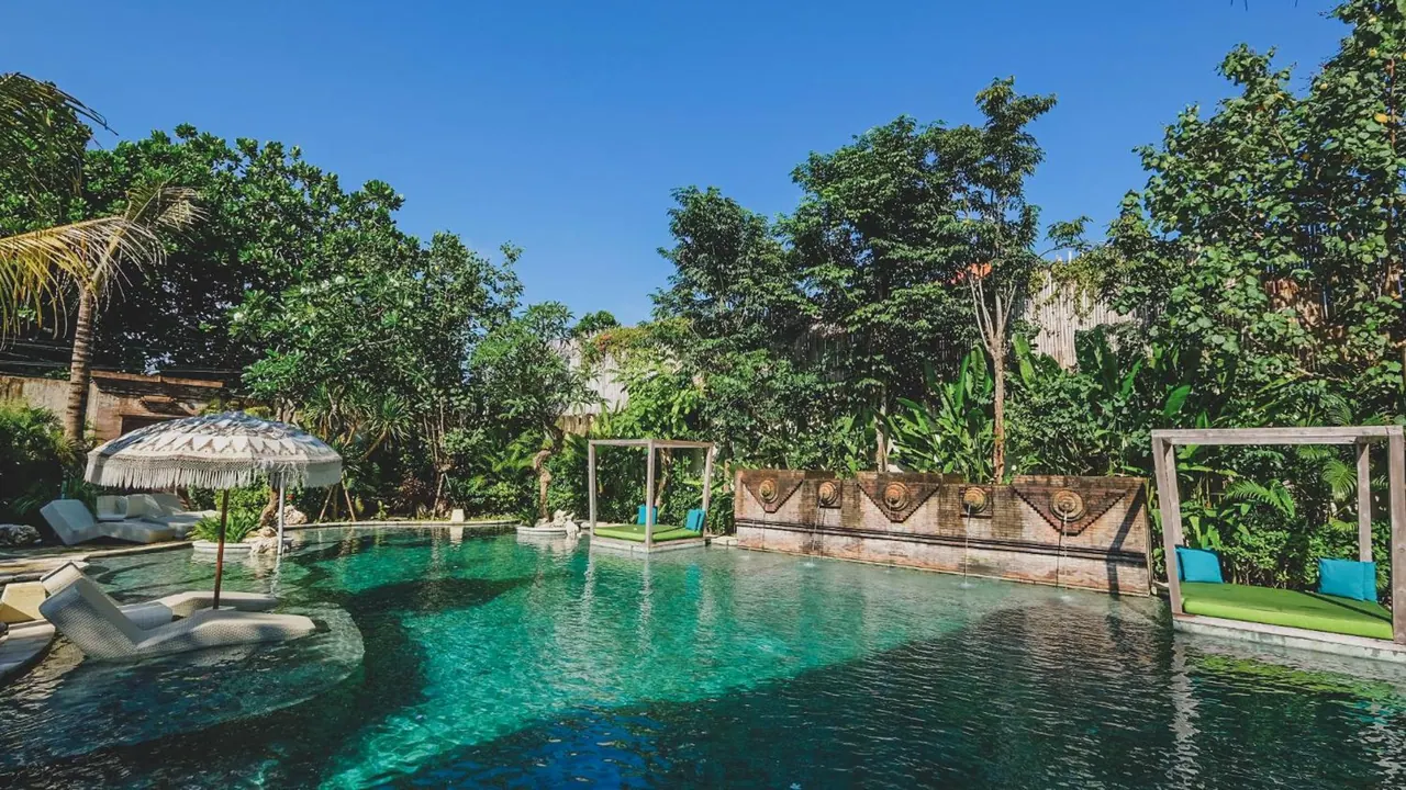 A large turquoise swimming pool with white loungers, a fringed parasol, green daybeds and a carved wall with water spouts, surrounded by tropical trees under a clear blue sky.