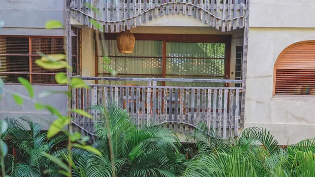 A wooden-railed balcony and arched wooden shutter on a cream building, with palm plants in the foreground and a rattan pendant lamp hanging beneath the balcony.