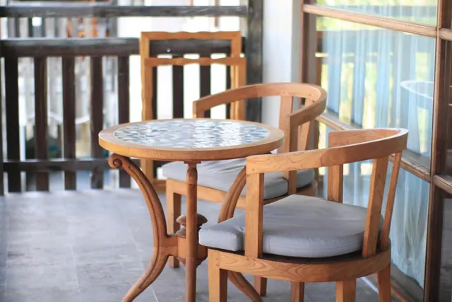 A round tiled-top table with two wooden armchairs and grey cushions sits on a balcony beside a wooden railing and a glass door with sheer curtains.