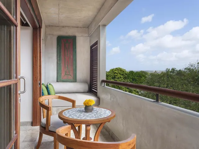 A covered balcony with a built-in bench, two wooden chairs and a mosaic-top table holding yellow flowers, overlooking treetops and sky.