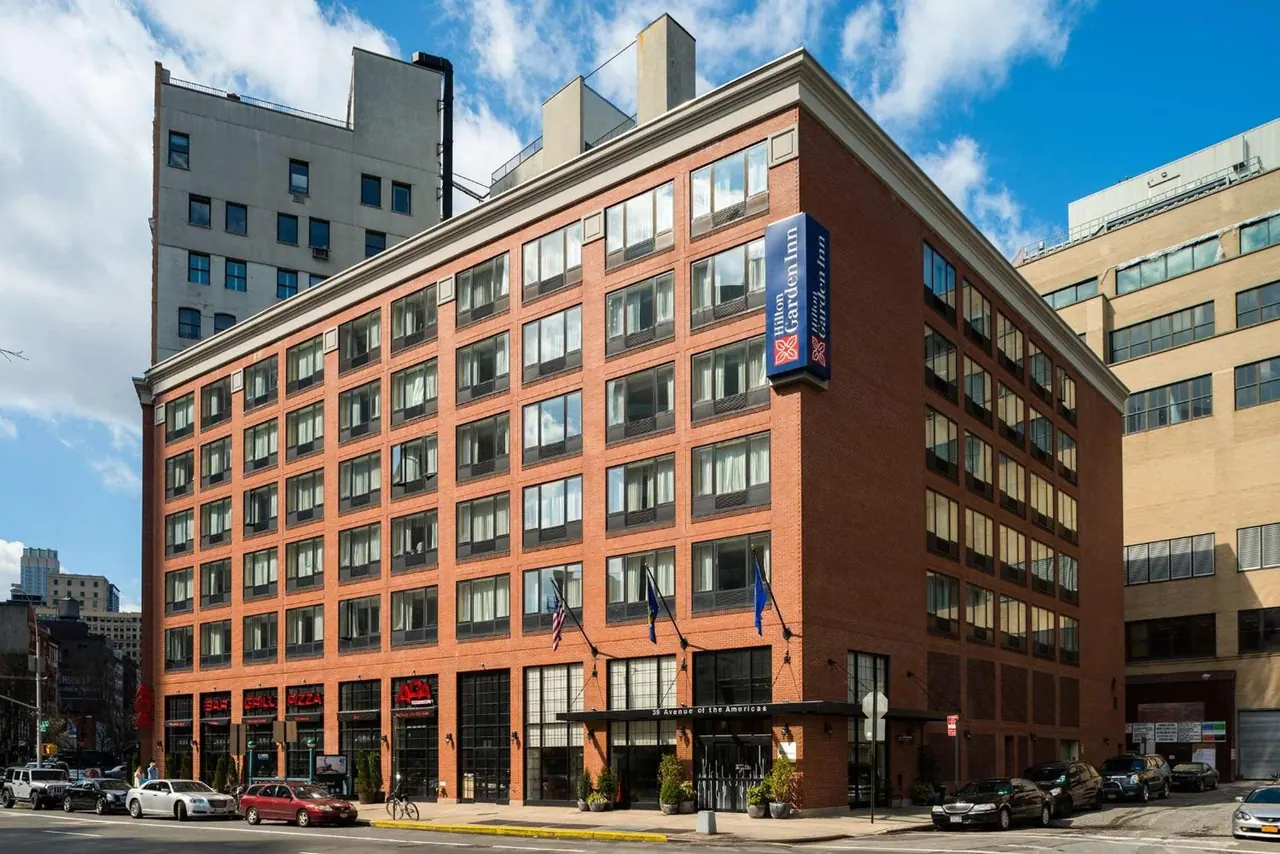 The exterior of a seven-storey red brick Hilton Garden Inn building stands on a city street with parked cars, a bicycle and taller buildings beside it under a partly cloudy sky.