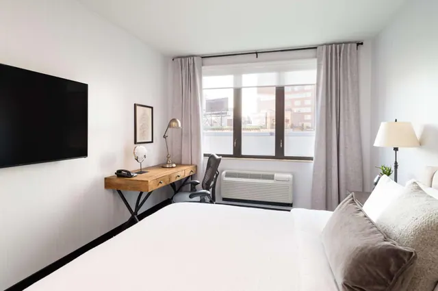 A bedroom with a king bed, a wall-mounted television, a wooden desk with a chair, and a window looking out onto nearby city buildings.