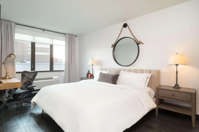 A bedroom with a made king bed, a round mirror hung on rope above the headboard, a desk and office chair by the window, and a wall-mounted air conditioner unit.