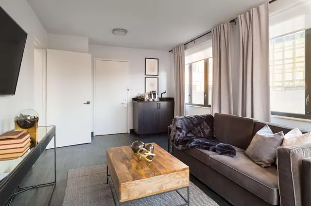A living area with a grey sofa, a wooden coffee table, a wall-mounted television, a coffee machine on a dark cabinet, and a window with curtains looking onto a nearby building.