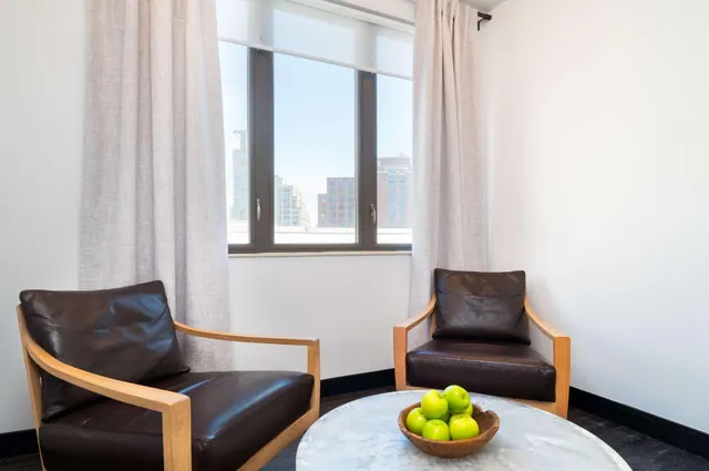 Two brown leather armchairs and a round marble table with a bowl of green apples sit by a window overlooking city buildings.