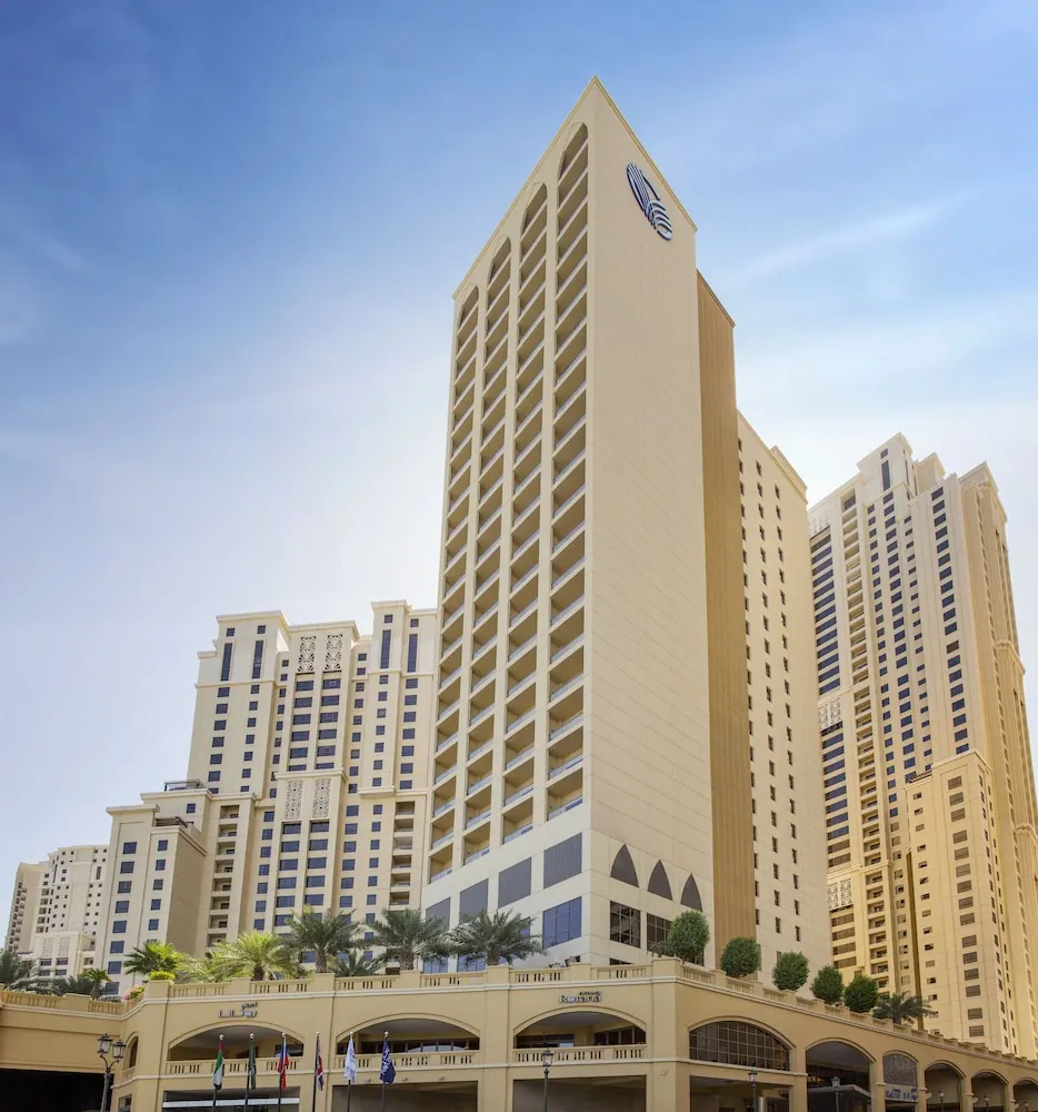 Low-angle view of a tall cream-coloured hotel tower with a blue logo near its top, flanked by residential towers, palm trees and flags at the base under a blue sky.