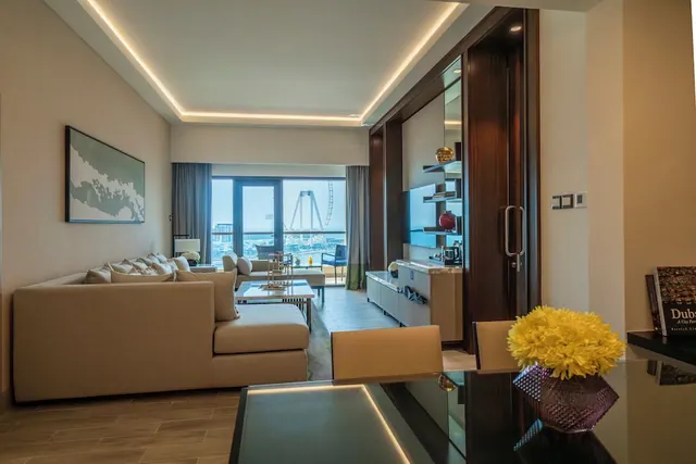 A living room with a beige sofa, coffee table and glass dining table, with a balcony door showing the Ain Dubai Ferris wheel outside.