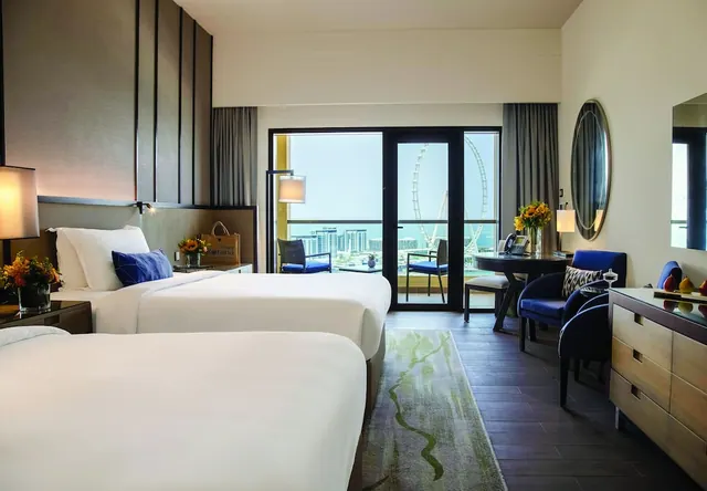 A twin-bedded hotel room with blue armchairs and a desk, with a balcony door looking out over the sea and the Ain Dubai ferris wheel.