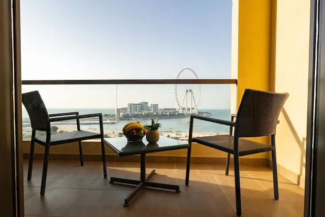 A balcony with two wicker chairs and a glass table holding a fruit bowl and plant, overlooking the sea, beach and the Ain Dubai ferris wheel.