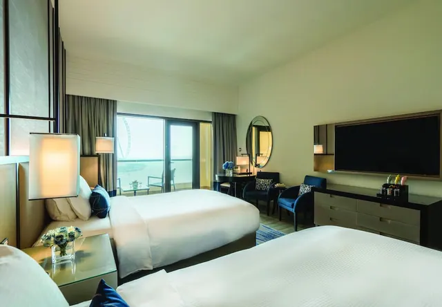 A hotel bedroom with two beds, a wall-mounted television, armchairs and sliding doors opening onto a balcony with a view of the Ain Dubai ferris wheel and the sea.