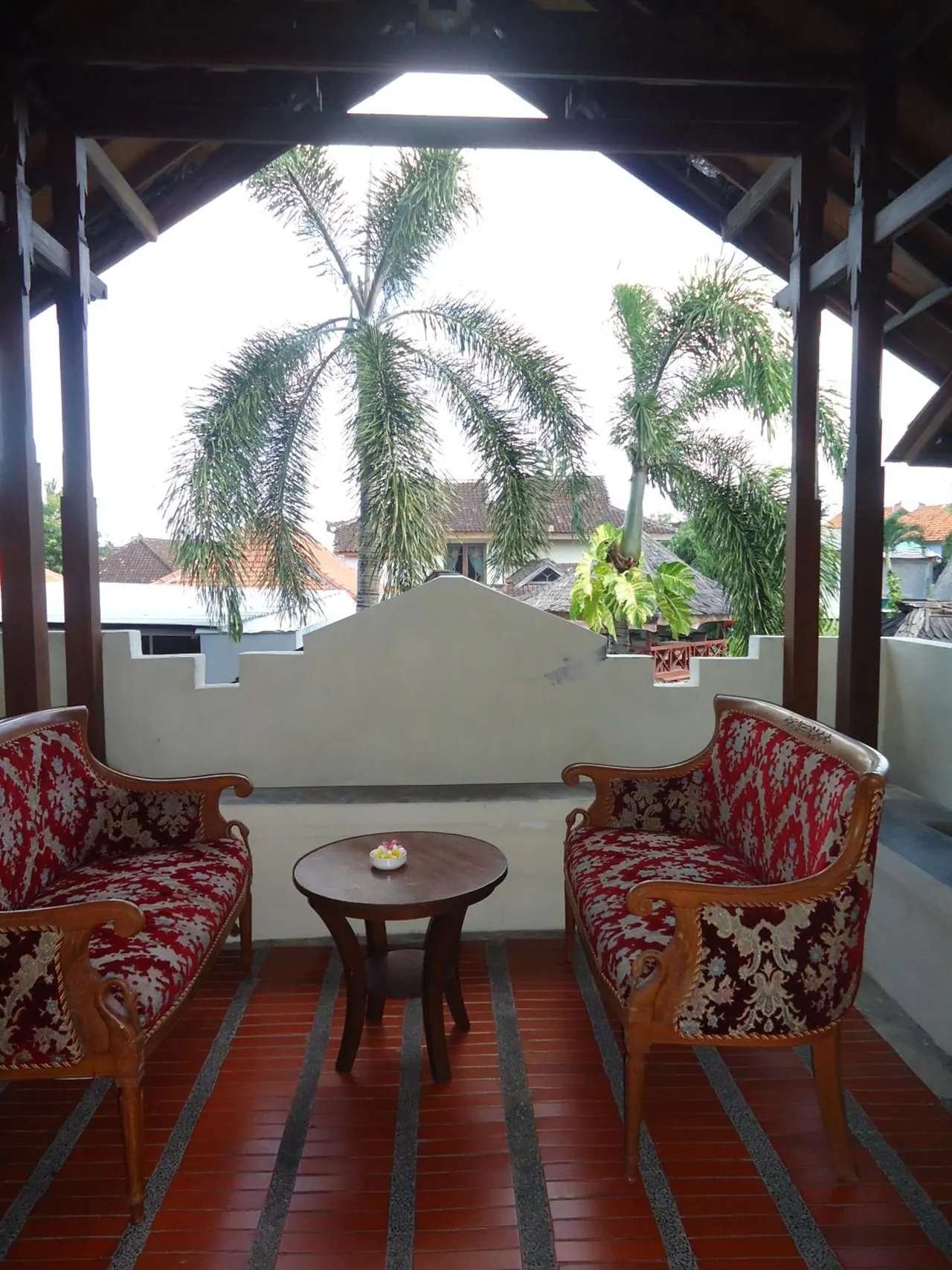 A covered terrace with two red upholstered armchairs and a small round wooden table, with palm trees and rooftops visible beyond the open side.