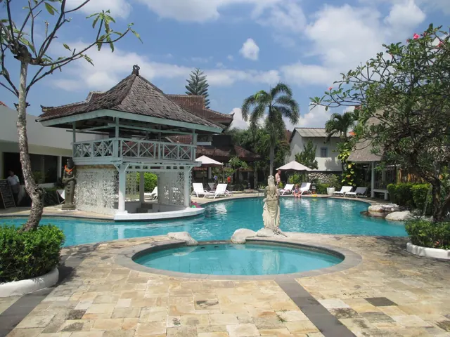 A large freeform swimming pool with a thatched-roof pool bar, a small round pool in the foreground, a statue, and sun loungers with parasols along the edge.