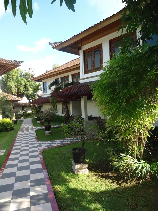 A two-storey white building with a tiled roof stands beside a checkered stone path, a lawn, potted plants and palm trees.