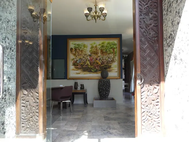 Carved wooden doors frame a lobby with a large painting on the back wall, a chandelier overhead, a dark armchair, a side table and a dark sculptural vase.