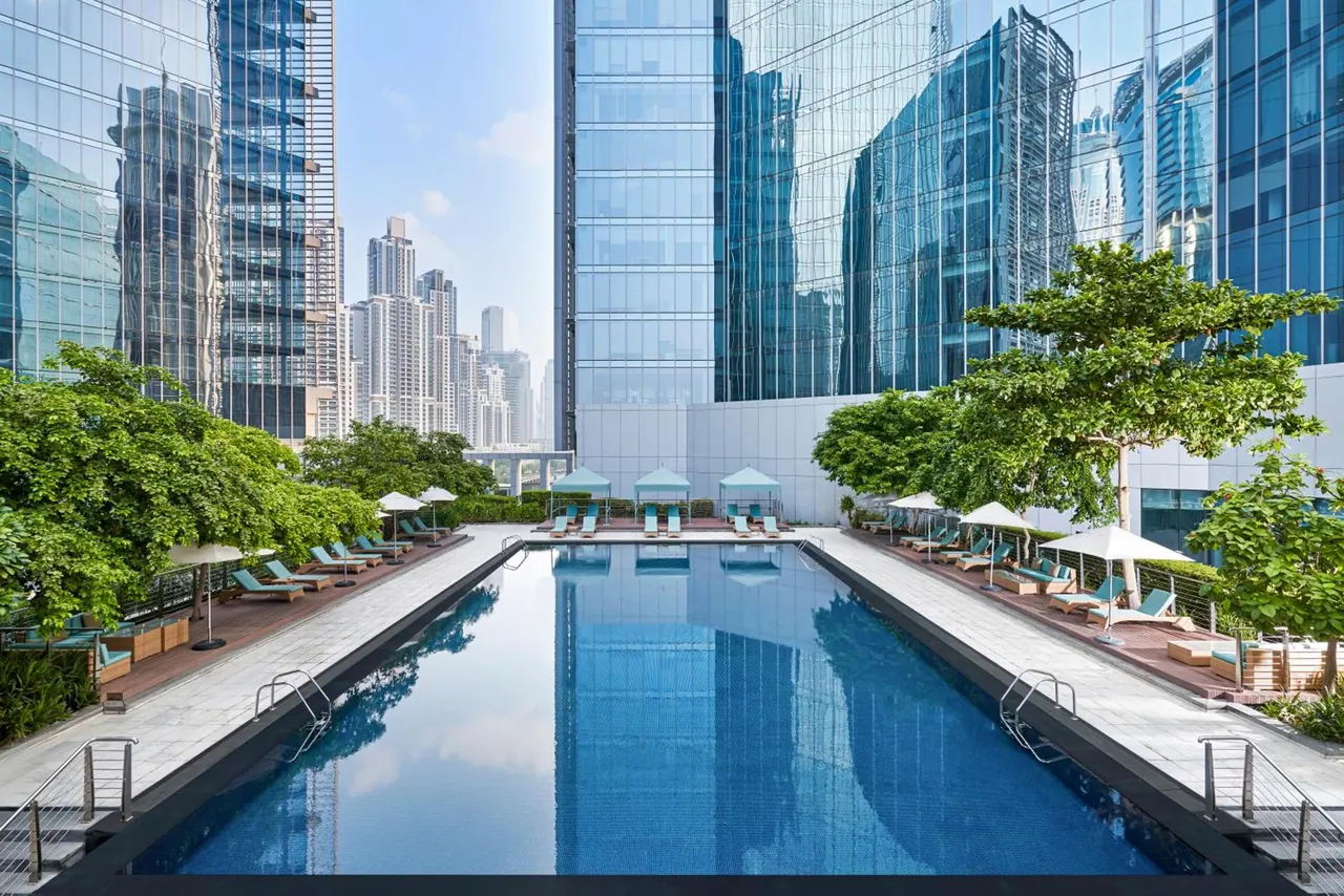 A long rectangular swimming pool runs between glass office towers, with teal sun loungers and white umbrellas along both sides and a distant city skyline beyond the trees.