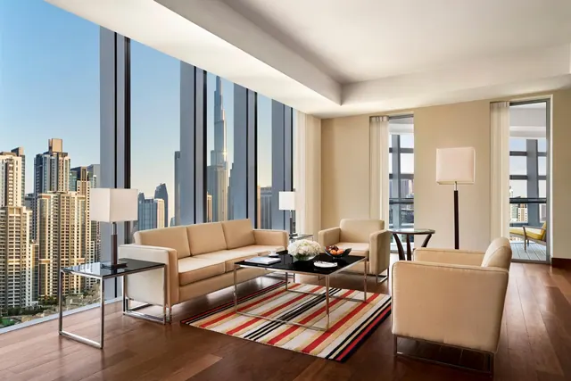 A living room with a sofa, armchairs and a striped rug beside floor-to-ceiling windows overlooking a city skyline with a tall tower, with an open door to a balcony.