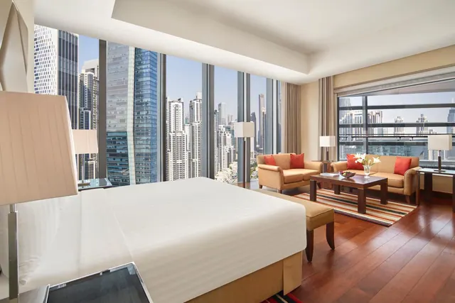 A hotel bedroom with a large bed beside floor-to-ceiling windows, with a seating area of two sofas and a coffee table, overlooking high-rise towers.