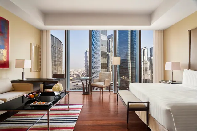 A hotel bedroom with a king bed, a sofa and armchair seating area, a work desk, and floor-to-ceiling windows overlooking high-rise towers.
