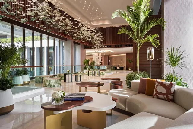 A hotel lobby with a cream sofa, low wooden and gold tables, potted palms and a suspended floral installation beneath a chandelier.