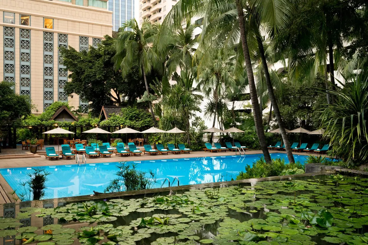 An outdoor pool with turquoise loungers and beige umbrellas, surrounded by palm trees, with a lily pond in the foreground.