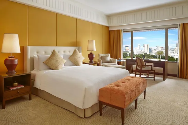 A bedroom with a king bed, two bedside lamps, an upholstered armchair, a desk by a large window, and a view of city buildings and trees.