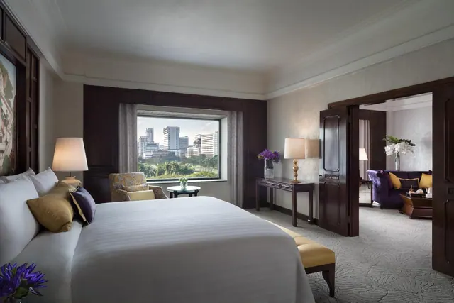 A bedroom with a made king bed, a window showing a city skyline, and an open doorway into an adjoining sitting room with a purple sofa.