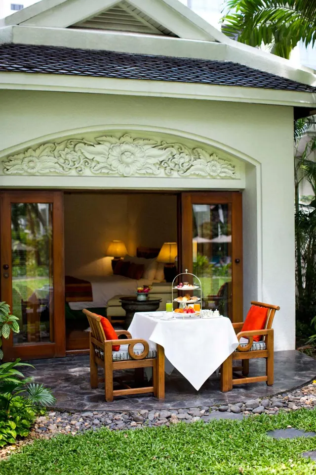 A set afternoon tea table with two wooden chairs and red cushions sits on a stone terrace outside a villa, with a bed visible through the open glass doors.